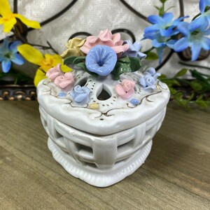 May include: White porcelain trinket box with a heart-shaped base and a lattice design. The lid is decorated with a cluster of pastel-colored flowers, including pink, blue, and yellow.