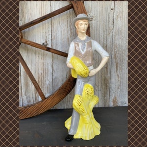 May include: A ceramic figurine of a person in a hat and vest, holding a bundle of yellow wheat. The figure is wearing a gray hat, vest, and pants, with a white shirt. The wheat is a bright yellow with small black dots. The figurine is standing in front of a wooden background.