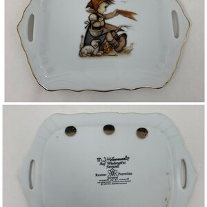Op de afbeelding: Een wit porseleinen dienblad met een gouden rand heeft een meisje met een rode capuchonjas en een mandje met kuikens. Het dienblad heeft twee handvatten en de achterkant is gemarkeerd met de tekst "M.J. Hummel, Auf Wiedersehn! Farewell, Reutter Germany Porzellan".