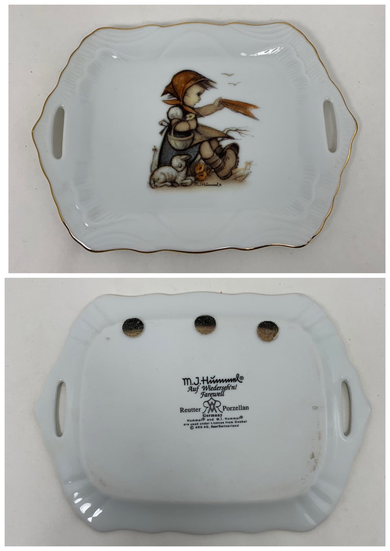 Op de afbeelding: Een wit porseleinen dienblad met een gouden rand heeft een meisje met een rode capuchonjas en een mandje met kuikens. Het dienblad heeft twee handvatten en de achterkant is gemarkeerd met de tekst "M.J. Hummel, Auf Wiedersehn! Farewell, Reutter Germany Porzellan".
