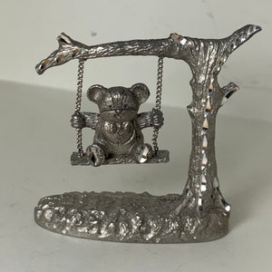 May include: A silver pewter figurine of a teddy bear on a swing. The swing is attached to a tree branch with a base.