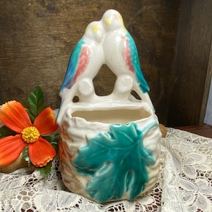 May include: A white ceramic wall planter shaped like a bird's nest with two blue, yellow, and pink birds perched on the rim. The planter has a green leaf design on the front.