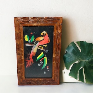 Peut inclure: Une &oelig;uvre d'art encadr&eacute;e repr&eacute;sentant un oiseau color&eacute; avec des plumes rouges, jaunes et vertes, peint sur un fond noir. Le cadre est en bois avec une finition rustique.