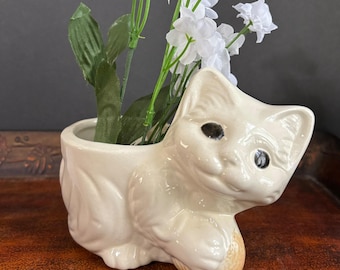 May include: A cream-colored ceramic cat planter holding a ball, with black eyes and a smiling expression. The planter is filled with white flowers and green leaves, creating a decorative floral arrangement.