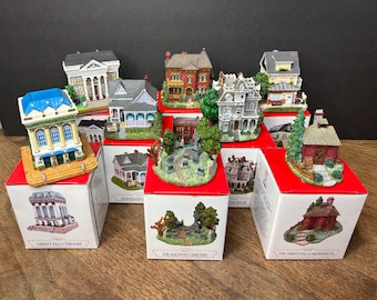 May include: A collection of miniature houses and buildings, each with intricate details and vibrant colors. The set includes a theater, cemetery, and various homes, all displayed on red and white boxes.