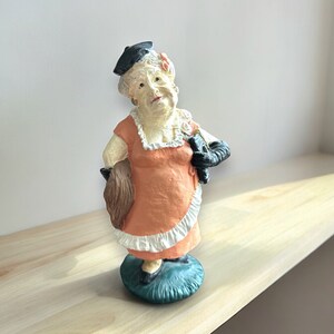 May include: A figurine of an older woman wearing a black hat, orange dress, and black gloves. She is holding a brown object in her left hand and has a whimsical expression on her face.