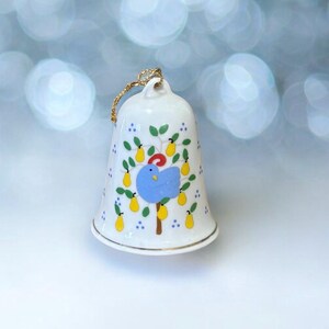 Puede incluir: Un adorno de campana de porcelana blanca con ribete dorado. La campana está decorada con un pájaro azul posado en una rama de árbol con peras amarillas. El árbol está rodeado de hojas verdes y puntos blancos.