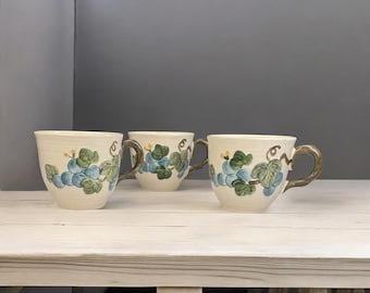 May include: Three white ceramic mugs with a grapevine design. The mugs have brown handles and are decorated with green leaves and blue grapes.