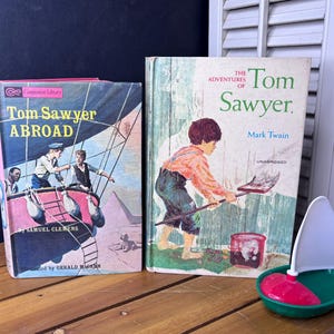 Op de afbeelding: Twee hardcover boeken getiteld Tom Sawyer Abroad van Samuel Clemens en The Adventures of Tom Sawyer van Mark Twain, met geïllustreerde covers met personages in een heteluchtballon en een jongen met een schep, naast een klein plastic speelgoedzeilbootje.
