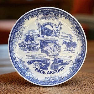 May include: A white and blue decorative plate featuring illustrations of landmarks from Page, Arizona, including Rainbow Bridge, Tower Butte, and Glen Canyon Dam. The plate has a detailed floral border and the text "PAGE. ARIZONA".