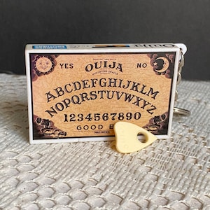 Può includere: Un tabellone Ouija in stile vintage con un planchette. Il tabellone è bianco e nero con lettere dorate e presenta le lettere dell'alfabeto, i numeri da 1 a 9 e lo 0, e le parole "YES", "NO", "GOODBYE" e "OUIJA".