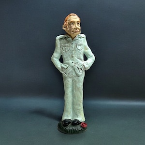 May include: A ceramic figurine of a person wearing a light blue shirt and pants. The figurine is standing on a green base with a red flower.