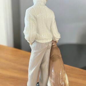May include: A porcelain figurine of a boy in a white sweater and gray pants standing with a brown and white dog. The boy is looking away from the camera and the dog is sitting with its head resting on the boy's leg.