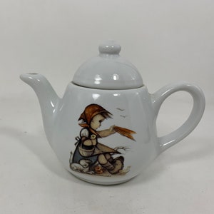 Op de afbeelding: Witte porseleinen theepot met een deksel, met een handgeschilderde illustratie van een meisje met een rode kap en twee konijnen. Het meisje houdt een stuk stof vast en er vliegen vogels in de achtergrond.