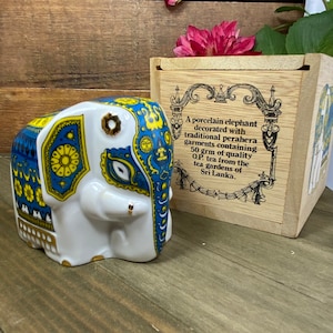 Puede incluir: Una figurita de elefante de porcelana blanca decorada con motivos tradicionales de perahera de Sri Lanka en azul y amarillo. El elefante está en una caja con una etiqueta que dice "Un elefante de porcelana decorado con motivos tradicionales de perahera que contiene 50 grm de té O.P. de calidad de los jardines de té de Sri Lanka."