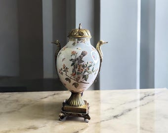 May include: A white ceramic vase with gold accents and a floral design featuring birds. The vase has a round body and a decorative base.