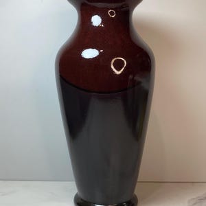 May include: A tall, glossy ceramic vase featuring a two-tone design with a dark mahogany brown upper section and a deep black lower section. The vase has a flared rim, a slender neck, and a tapered body that widens toward the center before narrowing.
