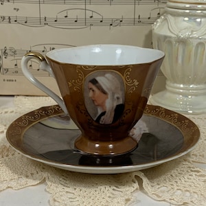 Pode incluir: Um conjunto de chávena e pires de chá castanho e dourado com um retrato de uma mulher com um chapéu branco. A chávena e o pires têm uma borda dourada e um desenho floral.