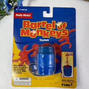 May include: A blue plastic barrel keychain with a red monkey hanging out of it. The packaging says "Barrel of Monkeys" and "Really Works!"  The item number is #537-0.