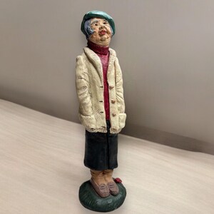 May include: A ceramic figurine of a woman wearing a green hat, a cream-colored jacket, a red sweater, and black pants. She is standing on a green base.