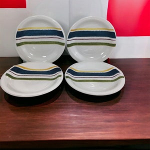 May include: Four white ceramic bowls with a striped design in shades of blue, green, yellow, and brown. The bowls are arranged in two rows on a wooden table.