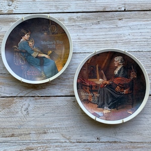 Può includere: Due piatti in ceramica bianca con bordo dorato, che presentano illustrazioni di Norman Rockwell. I piatti raffigurano scene di madri e figli. Il piatto superiore mostra una madre che legge al suo bambino vicino a un camino, con il testo "Mother's Day 1978". Il piatto inferiore mostra una madre che cuce mentre guarda un quadro incorniciato, con il testo "Mother's Day 1979".