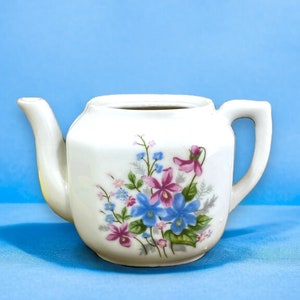 May include: A white teapot with a floral design featuring pink, blue, and purple flowers. The teapot has a spout and a handle.