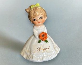 May include: A porcelain figurine of a girl wearing a white dress and holding an orange flower. The girl has blonde hair with a green bow and the word "August" is written on the base of the figurine.