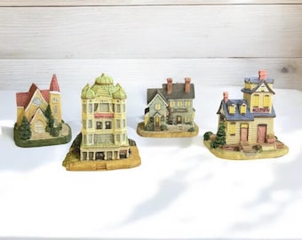 May include: Four miniature ceramic buildings, including a church, a multi-story building, and two houses. The buildings are painted in various colors, such as green, yellow, red, and blue. Each building is set on a base.