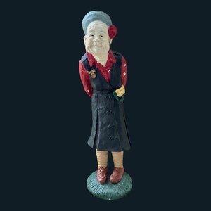 May include: A ceramic figurine of a woman with a red flower in her hair, wearing a black dress with a red and white patterned shirt, and holding a yellow flower. The figurine is standing on a green base.