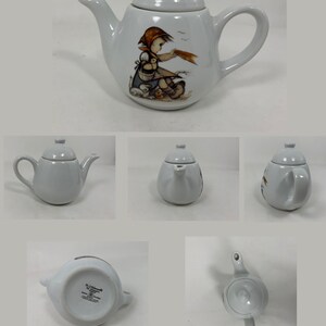 Op de afbeelding: Witte porseleinen theepot met een ontwerp van een meisje en vogels. De theepot heeft een deksel en een tuit. Op de bodem van de theepot staat in bruine letters "Hummel by Goebel Made in Germany".