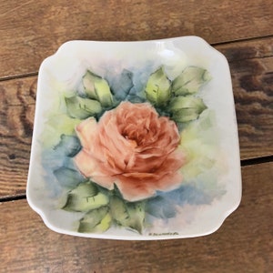 Pode incluir: Um prato quadrado de porcelana branca com um desenho pintado à mão de uma única rosa cor de pêssego com folhas verdes. O prato é assinado "H. Schwarzenbach" no canto inferior direito.