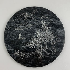 Puede incluir: Un posavasos redondo de mármol negro con un diseño grabado en blanco de un paisaje con árboles, montañas y un barco. El posavasos tiene los caracteres chinos "石  砚" grabados en la parte superior central.