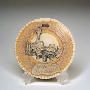 Puede incluir: Un pequeño plato de cerámica con una imagen en 3D en relieve de la Ciudad Vieja de Jerusalén. El plato tiene un esmalte marrón y blanco con un anillo marrón alrededor del borde. El plato está sobre un soporte blanco.
