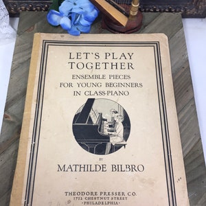Puede incluir: Portada de libro vintage con el título "Let's Play Together" en texto negro. La portada presenta una ilustración en blanco y negro de niños tocando un piano. El libro es de Mathilde Bilbro y publicado por Theodore Presser Co. en Filadelfia.