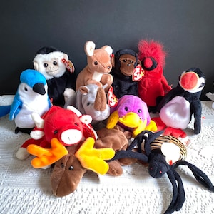 May include: A collection of 10 plush toys, including a blue jay, a red and white reindeer, a brown moose, a purple octopus, a brown rhino, a brown deer, a monkey, a red bird, a white and black penguin, and a black and yellow spider.