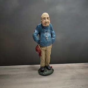 May include: A figurine of a bald man wearing a blue sweater with a whistle on a chain around his neck. He is holding a red baseball cap in his left hand and is standing on a green base.