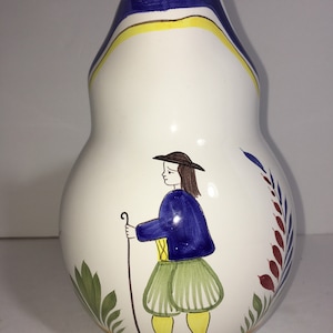 May include: A white ceramic vase with a pear-shaped body and a blue and yellow striped neck. The vase features a hand-painted design of a person in a blue jacket and green pants holding a walking stick. The person is standing in front of a green and red floral design.
