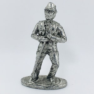 May include: A silver-colored metal figurine of a person wearing a hat and holding a stick. The figurine is standing on a small base.