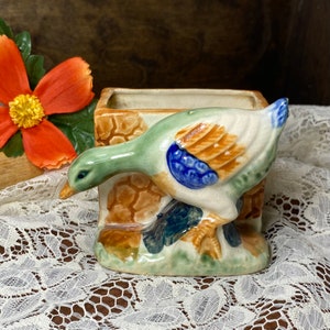 May include: A ceramic planter shaped like a duck with a brown, green, blue, and white glaze. The duck is perched on a brown, green, and white glazed base.