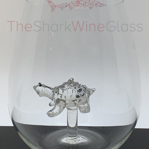 May include: A clear glass wine glass with a small, clear glass turtle figurine attached to the stem. The glass has the text "TheSharkWineGlass" printed on the side.