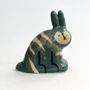Może przedstawiać: Ceramiczna figurka królika w pozycji siedzącej. Królik jest głównie ciemnozielony z różowymi paskami i beżową twarzą. Uszy są wyprostowane, a oczy to małe czarne kropki. Figurka ma fantazyjny, artystyczny design.