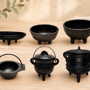 May include: A collection of six black cast iron vessels including three open bowls with tripod legs and three lidded pots with metal handles. The items vary in shape and size, featuring textured surfaces and sturdy construction for tabletop use.