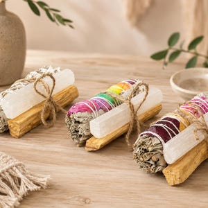 May include: Three bundles of dried sage and palo santo wood sticks tied together with twine. Each bundle includes a white selenite crystal wand. One bundle features rainbow-colored flower petals, another has pink petals, and the third has red and yellow petals.