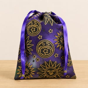 May include: A purple drawstring pouch featuring a repeating gold-colored print of suns, moons, and stars. The bag is closed with two vibrant purple satin ribbons that hang down the front, each finished with a small metallic bead at the end.