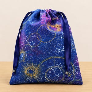 May include: A drawstring pouch featuring a deep blue fabric printed with a celestial pattern of white constellations, golden suns, and purple nebula clouds. The bag is cinched at the top with two dark blue satin ribbons and silver-toned beads.