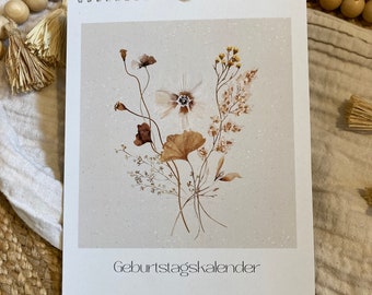 May include: A spiral-bound calendar featuring a watercolor illustration of dried flowers in shades of brown, beige, and white. The text "Geburtstagskalender" is printed at the bottom, with the words "mein goldstück" below it. The calendar is displayed on a neutral-toned surface.