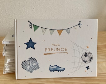 May include: A white hardcover book with a soccer theme. The cover features a soccer ball, cleats, goalie gloves, and a banner with the text "meine FREUNDE". The design includes blue and gold accents, with a stack of similar books visible.