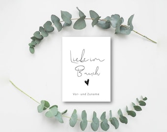 May include: A white card with black calligraphy that reads "Liebe im Bauch" with a small black heart below the text. The text is followed by "Vor- und Zuname". The card is surrounded by eucalyptus branches.