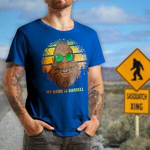 May include: A blue t-shirt with a retro-style graphic of a smiling sasquatch wearing sunglasses. The text "MY NAME IS DARRELL" is printed below the graphic.